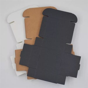 K&ouml;nnte beinhalten: Drei Pappkartons in verschiedenen Farben: Wei&szlig;, Braun und Schwarz. Die Kartons sind flach gefaltet und haben ein schlichtes Design.