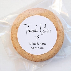 Pode incluir: Um biscoito redondo em uma embalagem plástica transparente. O biscoito tem um rótulo circular branco com as palavras "Thank You" em fonte cursiva, um símbolo de coração e os nomes "Mike & Kate" com a data "08.06.2028".
