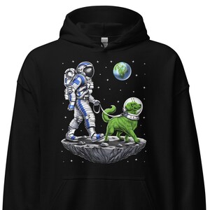 Puede incluir: Una sudadera con capucha negra con un gráfico de un astronauta paseando a un perro alienígena verde por la luna. El astronauta lleva un traje espacial blanco con detalles azules. El perro lleva un collar azul y blanco. El fondo es un espacio negro con estrellas y un planeta.