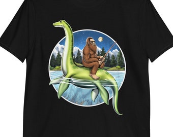 Puede incluir: Una camiseta negra con un gráfico de un monstruo marino prehistórico verde con un sasquatch marrón montando en su espalda y sosteniendo una caña de pescar. El sasquatch lleva una bandana roja.