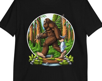 Könnte beinhalten: Schwarzes T-Shirt mit einem Aufdruck von Bigfoot, der einen Fisch und eine Angelrute in einer Waldlandschaft hält.