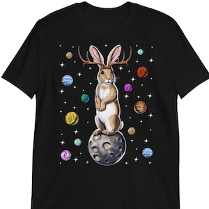 Pode incluir: Camiseta preta com uma ilustração fantástica de um coelho com chifres, em pé sobre uma lua, rodeado por planetas e estrelas. O design tem um tema espacial.