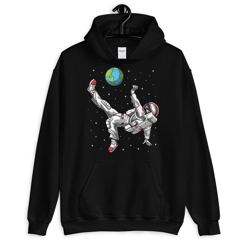 May include: Black hooded sweatshirt with a graphic of an astronaut kicking a soccer ball that is the Earth. The astronaut is wearing a white spacesuit with red accents. The background is a black space with white stars.