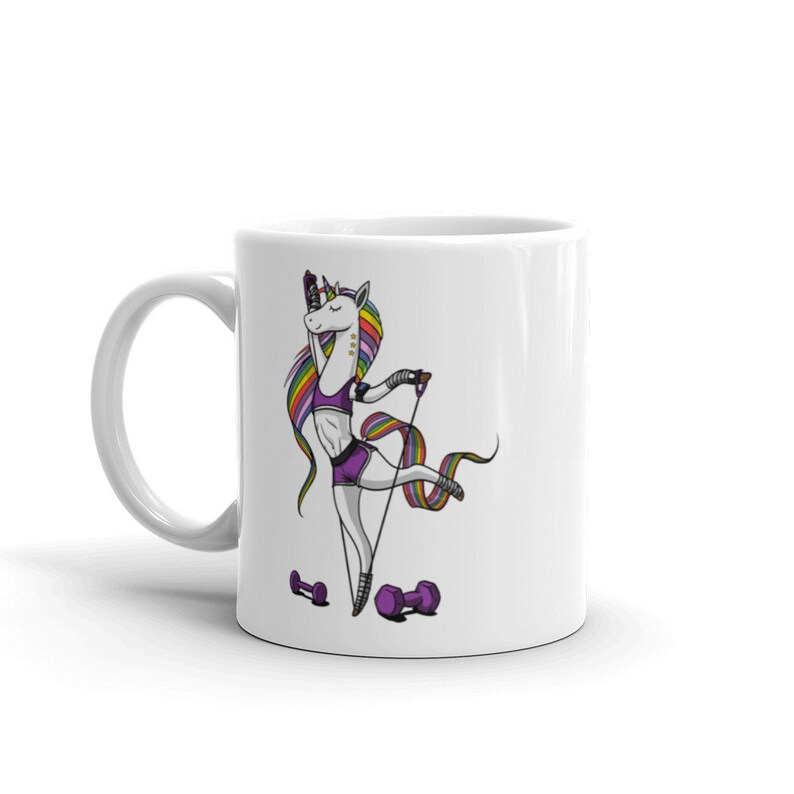 Op de afbeelding: Een witte keramische koffiemok met een kleurrijke illustratie van een eenhoorn. De eenhoorn draagt een paarse sportbeha en shorts en houdt een weerstandsband vast. De eenhoorn heeft een regenboogmanen en een regenboogstaart. Er zijn twee paarse dumbbells aan de onderkant van de mok.
