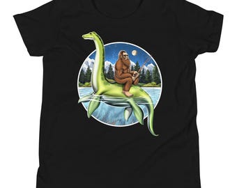 Puede incluir: Una camiseta negra con un gráfico de un Bigfoot montando un monstruo marino verde mientras pesca. El gráfico está sobre un fondo azul con una luna y estrellas.