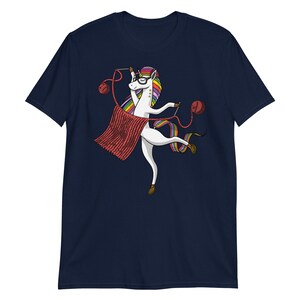 Puede incluir: Una camiseta azul marino con un unicornio blanco que lleva gafas y teje una bufanda roja. El unicornio tiene una melena y una cola arco&iacute;ris.