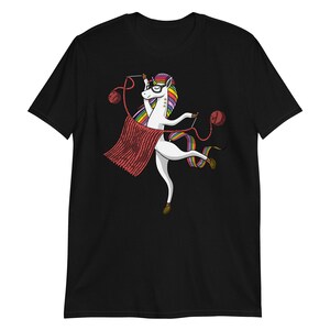Puede incluir: Una camiseta negra con un unicornio blanco que lleva gafas y teje una bufanda roja. El unicornio tiene una melena y una cola arco&iacute;ris.