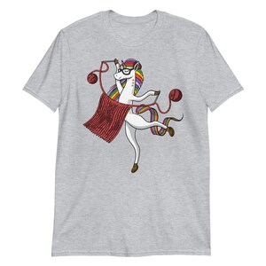 Puede incluir: Una camiseta gris con un unicornio blanco que lleva gafas y teje una bufanda roja. El unicornio tiene una melena y una cola arco&iacute;ris.