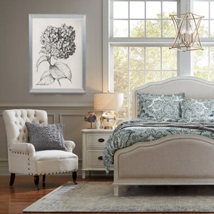 May include: A white upholstered bed with a headboard and footboard, featuring a gray and green patterned duvet and pillows. A white armchair with tufted upholstery and a gray throw pillow sits in the foreground. A black and white framed print of a hydrangea flower hangs on the wall.