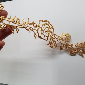 May include: A gold-coloured, floral-themed decorative element. The design features intricate cut-out roses and leaves, creating a delicate and ornate appearance. The piece is likely made of metal and has a shiny finish.