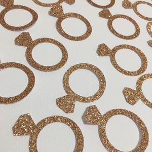 Puede incluir: Una colecci&oacute;n de confeti en forma de anillo de color dorado con purpurina. El confeti es perfecto para a&ntilde;adir un toque de brillo a la decoraci&oacute;n de bodas, despedidas de soltera u otros eventos especiales.