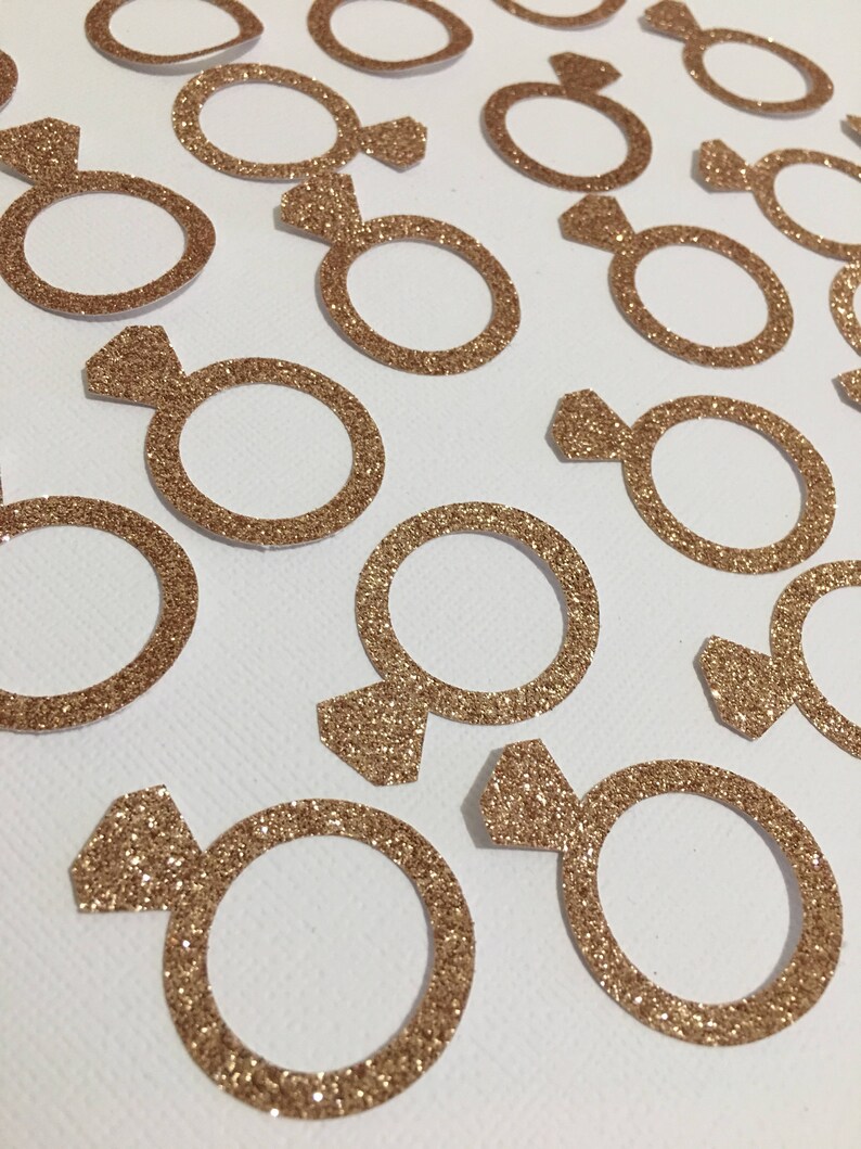 Puede incluir: Una colecci&oacute;n de confeti en forma de anillo de color dorado con purpurina. El confeti es perfecto para a&ntilde;adir un toque de brillo a la decoraci&oacute;n de bodas, despedidas de soltera u otros eventos especiales.