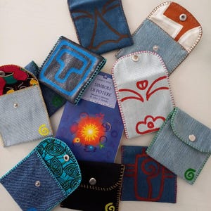 Può includere: Una collezione di piccole pochette in denim fatte a mano con vari disegni e colori. Alcune pochette presentano simboli dipinti, mentre altre hanno fodere in tessuto fantasia. È visibile anche un libro intitolato "Simboli di Potere".