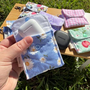 May include: A blue daisy-print pouch containing white wireless earbuds. Other small fabric pouches in various patterns and colours, including pink gingham, floral, and purple, are visible. The pouches are on a wooden surface, with green grass in the background.