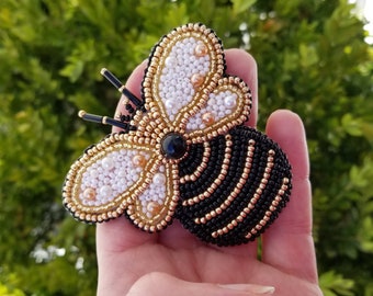 May include: Handmade beaded bee brooch. The bee features a black body with gold stripes, white and gold wings, and a black gem center. The brooch is a unique accessory.