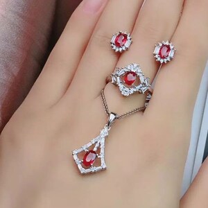 May include: A silver ring, necklace, and earrings set with red oval gemstones surrounded by smaller clear gemstones.