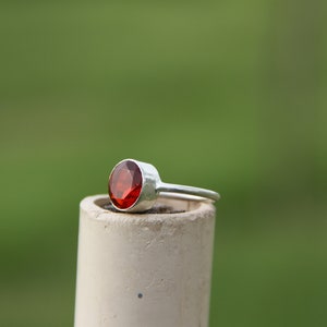 May include: A silver ring with a red gemstone set in the center. The ring is sitting on a white cylinder.