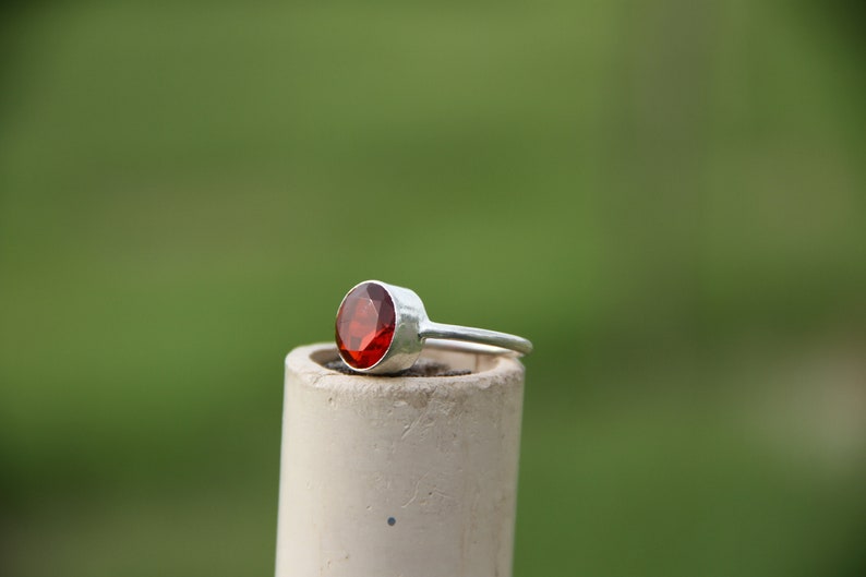 May include: A silver ring with a red gemstone set in the center. The ring is sitting on a white cylinder.