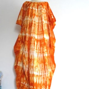 Peut inclure: Une robe caftan longue et ample présentant un motif tie-dye orange et blanc éclatant. Le vêtement possède des manches courtes et larges, une encolure arrondie et une silhouette fluide qui tombe des épaules jusqu'à l'ourlet.