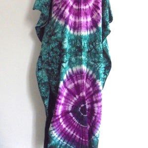 Könnte beinhalten: Ein langes, fließendes Batik-Kleid in Türkis-, Lila- und Weißtönen. Das Kleid hat ein großes, kreisförmiges Muster auf der Vorder- und Rückseite, einen quadratischen Ausschnitt und weite Ärmel. Das Kleid wird auf einer Schaufenquin präsentiert.