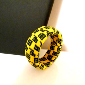 Könnte beinhalten: Ein gelbes Stoffarmband mit einem sich wiederholenden schwarzen Quadratmuster. Das Armband ist kreisförmig und scheint aus einem weichen Material zu bestehen. Der Hintergrund ist weiß und schwarz.