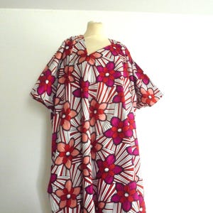Op de afbeelding: Een levendige jurk met bloemenprint en een V-hals. De jurk heeft een opvallend patroon van grote, gestileerde bloemen in rood, roze en wit, tegen een gestreepte achtergrond. De jurk wordt op een mannequin gepresenteerd.