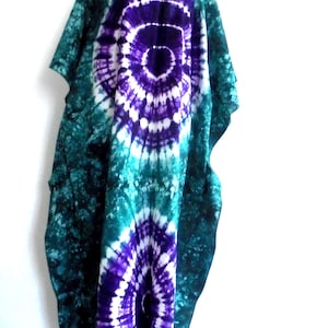Peut inclure: Un caftan tie-dye dans les tons de turquoise, de violet et de blanc. Le vêtement présente un grand motif en spirale central violet et blanc, avec un fond turquoise. Le caftan est présenté sur un mannequin sur fond blanc.