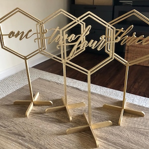 Puede incluir: Números de mesa geométricos dorados. Cada número se muestra en un marco hexagonal con una base en forma de cruz. Los números están escritos en cursiva. Los números son uno, dos, cuatro y tres.