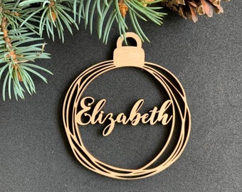 May include: A wooden Christmas ornament shaped like a circle with the name "Elizabeth" written in cursive inside. The ornament is on a dark gray background.