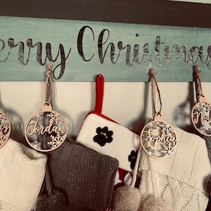 May include: A horizontal wooden sign painted light blue with the text Merry Christmas in black script. Hanging from the sign are circular wooden ornaments with snowflake cutouts and names, alongside white and gray knit stockings and a paw print stocking.