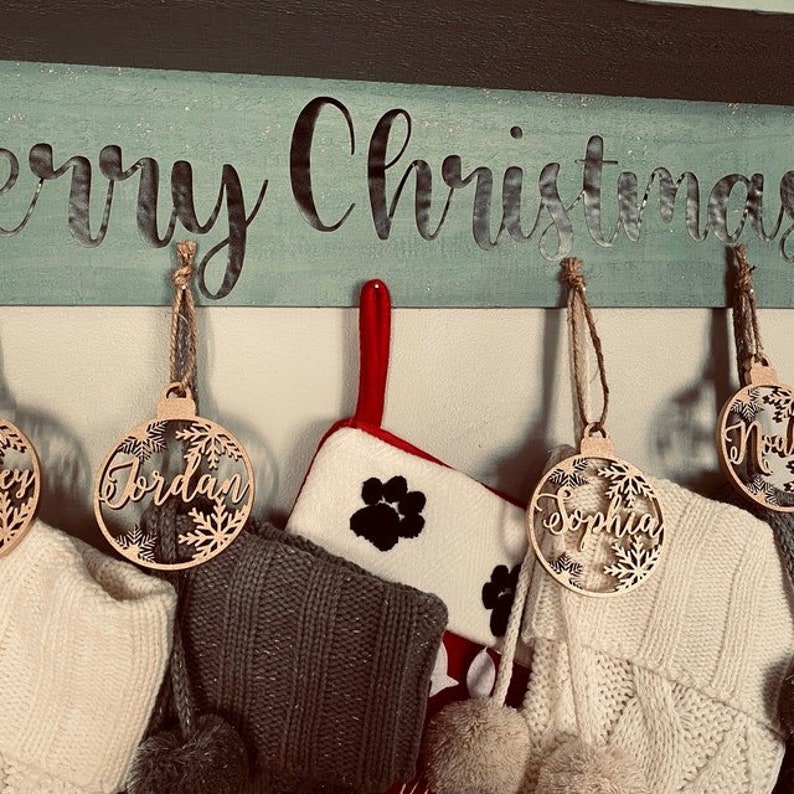 May include: A horizontal wooden sign painted light blue with the text Merry Christmas in black script. Hanging from the sign are circular wooden ornaments with snowflake cutouts and names, alongside white and gray knit stockings and a paw print stocking.