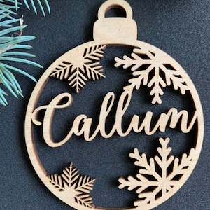 May include: A circular light brown wooden Christmas ornament featuring the name Callum in a cursive script font. The design includes three laser-cut snowflake patterns integrated into the frame, with a small loop at the top for hanging.