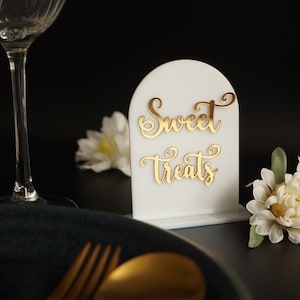 May include: White acrylic sign with a rounded top and gold lettering that reads "Sweet Treats". The sign is sitting on a black surface with a white flower in the background.