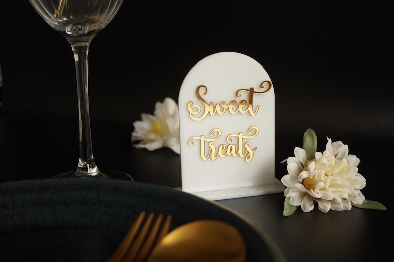 May include: White acrylic sign with a rounded top and gold lettering that reads "Sweet Treats". The sign is sitting on a black surface with a white flower in the background.