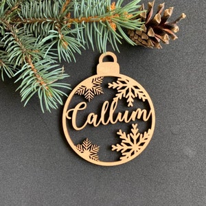 May include: A circular light brown wooden ornament featuring the name Callum in a cursive script font. The design includes cut-out snowflake patterns within the circular frame and a small loop at the top for hanging, presented with pine needles.