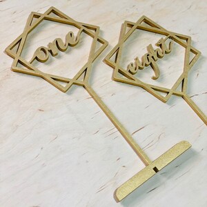 May include: Two gold table number holders with geometric designs. The numbers "one" and "eight" are cut out of the holders.