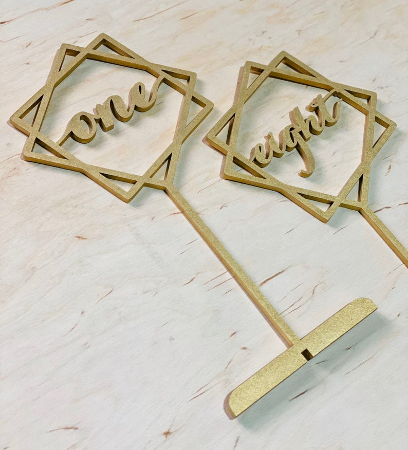 May include: Two gold table number holders with geometric designs. The numbers "one" and "eight" are cut out of the holders.