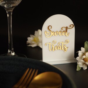 May include: White acrylic sign with a gold cursive inscription that reads "Sweet Treats". The sign is on a black surface with a white flower and a wine glass.