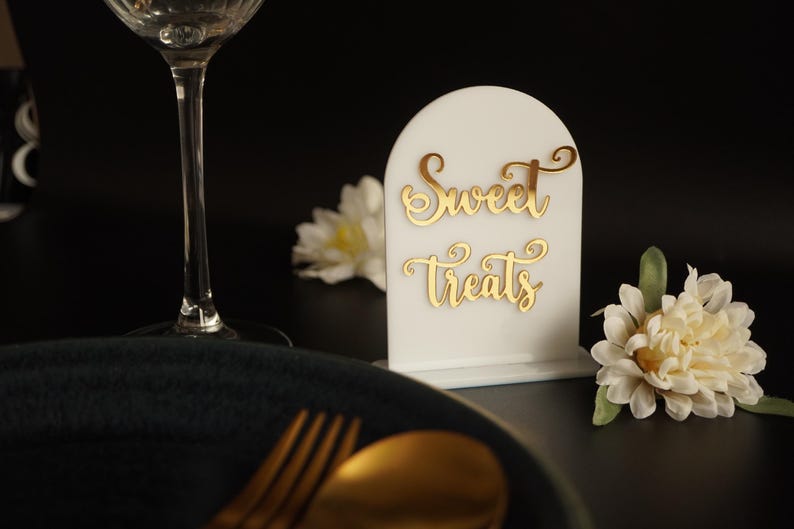 May include: White acrylic sign with a gold cursive inscription that reads "Sweet Treats". The sign is on a black surface with a white flower and a wine glass.