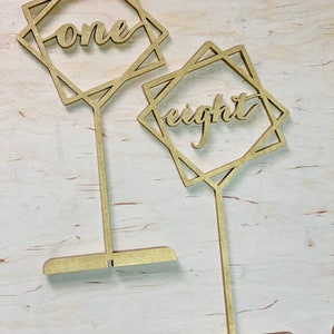 May include: Two gold wooden table numbers with geometric frames. The numbers "one" and "eight" are written in a cursive font.