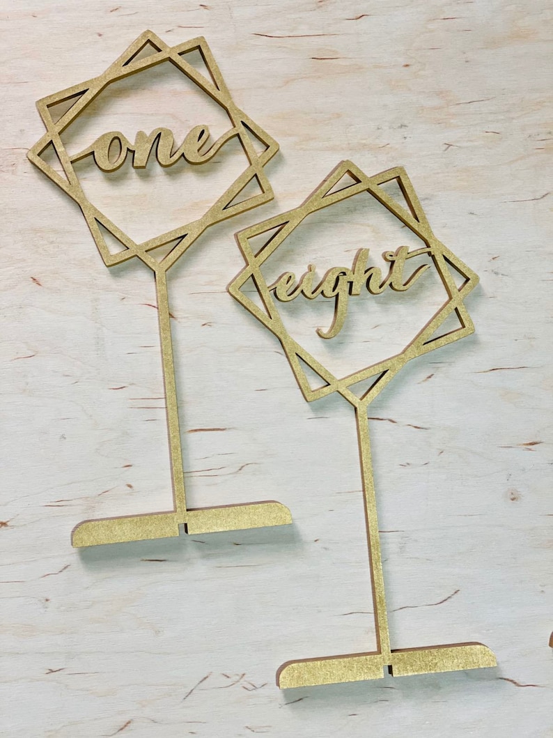 May include: Two gold wooden table numbers with geometric frames. The numbers "one" and "eight" are written in a cursive font.