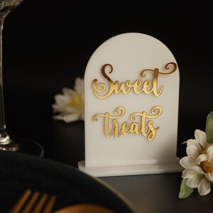 May include: White acrylic sign with a rounded top and gold lettering that reads "Sweet Treats". The sign is sitting on a table with a white flower in the foreground.