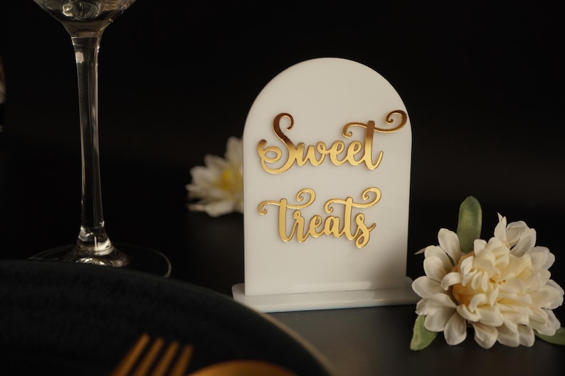 May include: White acrylic sign with a rounded top and gold lettering that reads "Sweet Treats". The sign is sitting on a table with a white flower in the foreground.