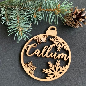 May include: A circular wooden ornament featuring the name Callum in a cursive script, surrounded by cut-out snowflake designs. The ornament has a small loop at the top for hanging and is positioned near a green pine branch and a brown pinecone.