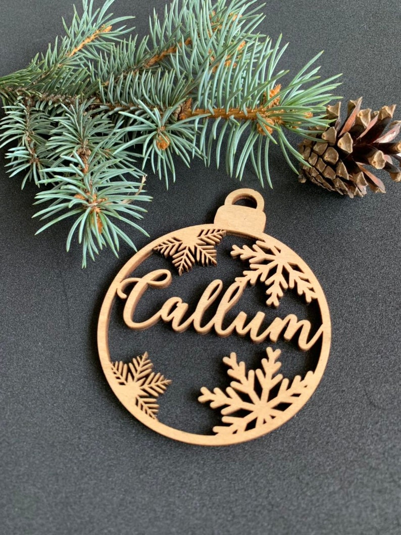 May include: A circular wooden ornament featuring the name Callum in a cursive script, surrounded by cut-out snowflake designs. The ornament has a small loop at the top for hanging and is positioned near a green pine branch and a brown pinecone.