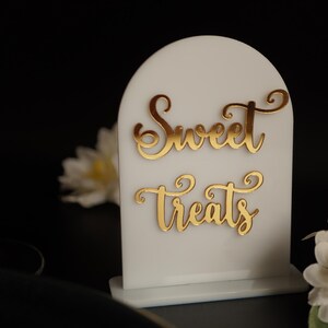 May include: White acrylic sign with a rounded top and a gold inscription that reads "Sweet Treats".