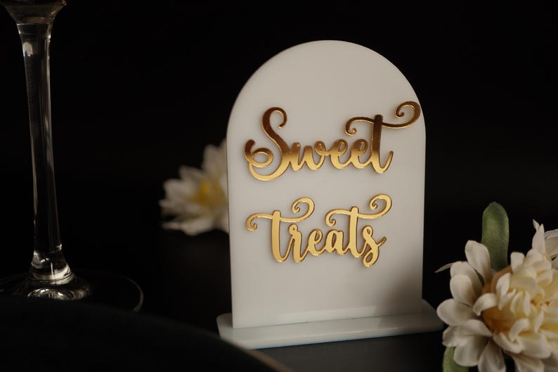 May include: White acrylic sign with a rounded top and a gold inscription that reads "Sweet Treats".