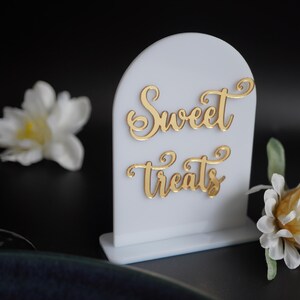 May include: White acrylic sign with a gold cursive inscription that reads "Sweet Treats". The sign is on a white base and has a rounded top.