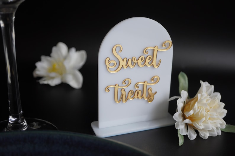May include: White acrylic sign with a gold cursive inscription that reads "Sweet Treats". The sign is on a white base and has a rounded top.