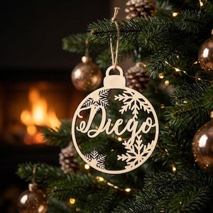 May include: A circular light-colored wooden Christmas ornament featuring a cutout design of snowflakes and the name Diego in a cursive script font. The ornament is suspended by a thin twine string from a branch of a green pine tree.